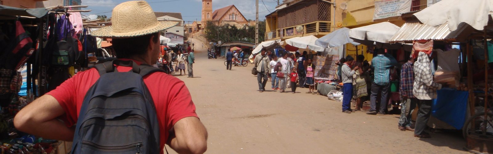 Peace Corps header image of a man in a red shirt and straw hat walking down a dirt road filled with vendors and people